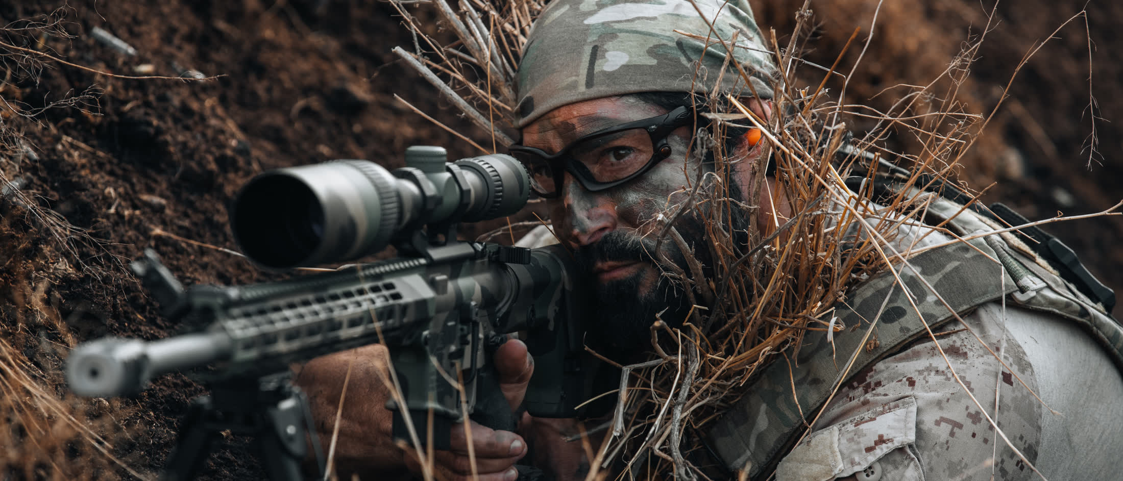 Operator prone in dry grass sighting a scoped rifle on a hillside
